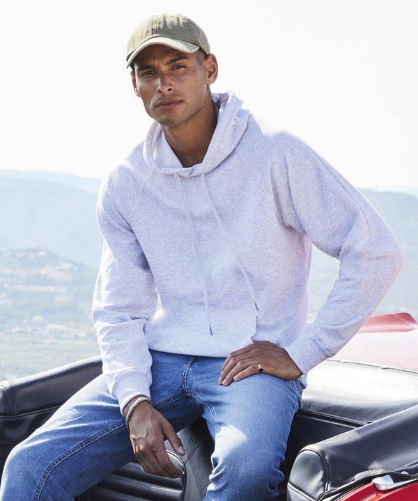 A person wearing a light gray SS925 hoodie from Fruit of the Loom, blue jeans, and a beige cap sits on a car trunk with mountains in the background.