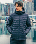 A person wearing a dark Stormtech ST013 puffer jacket stands near a body of water with a cityscape and boats in the background.