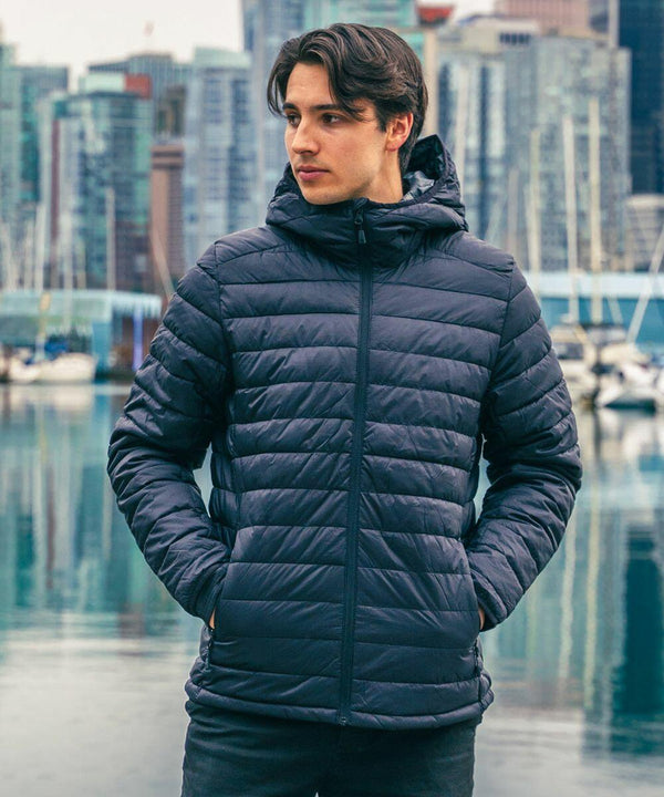 A person wearing a dark Stormtech ST013 puffer jacket stands near a body of water with a cityscape and boats in the background.