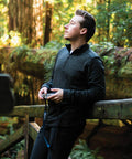 A person in a dark Stormtech ST021 pullover stands outdoors, holding a camera, and looking up thoughtfully with trees in the background.
