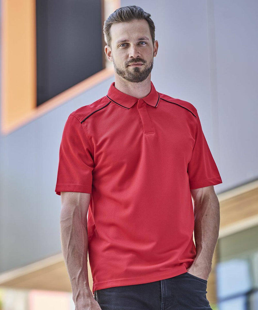 A man with short hair and a beard is wearing a red Stormtech ST078 polo shirt and standing outdoors in front of a modern building.