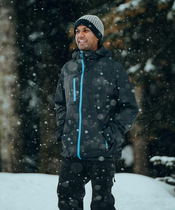 A person in a black Stormtech ST079 jacket and beanie stands smiling outdoors in a snowy environment. Snowflakes are falling around them.