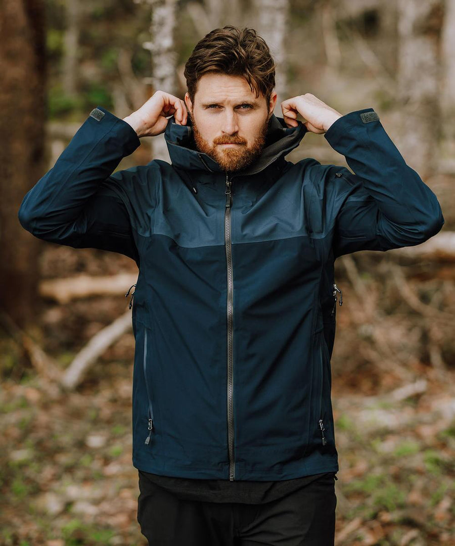 A bearded man stands outdoors in front of trees, wearing the Stormtech Vertex Stormshell dark blue hooded jacket and adjusting its collar with both hands.