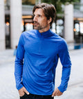 A man with brown hair and a beard stands outdoors on a city street, wearing the Stormtech Milano 1/4-zip jumper in blue and jeans, hands in his pockets, looking to the left.