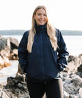 A woman with long blonde hair stands outdoors by rocky water, smiling with her hands in the pockets of a navy Women's Scirocco lightweight shell from Stormtech. The background features calm water and distant trees.
