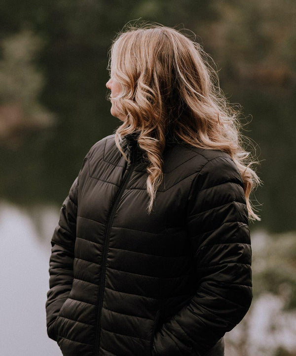 A person with long, wavy hair wearing a Stormtech ST144 black quilted jacket stands outdoors with a blurred background of water and trees.