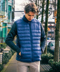A person wearing a blue Stormtech ST158 quilted vest and black long-sleeve shirt stands on a sidewalk next to buildings and trees, with hands in pockets.