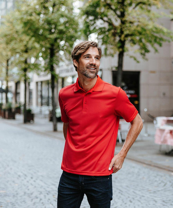 A man wearing a red Stormtech ST172 stands on a cobblestone street flanked by trees and buildings, looking to the side and smiling.