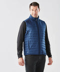 A man stands wearing a blue quilted Stormtech ST174 vest over a black long-sleeve shirt and black pants, against a plain white background.