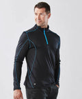 A man is wearing a Stormtech ST177 black long-sleeve athletic shirt with blue piping and a front zip. He stands with his hands in his pockets, looking to the side.