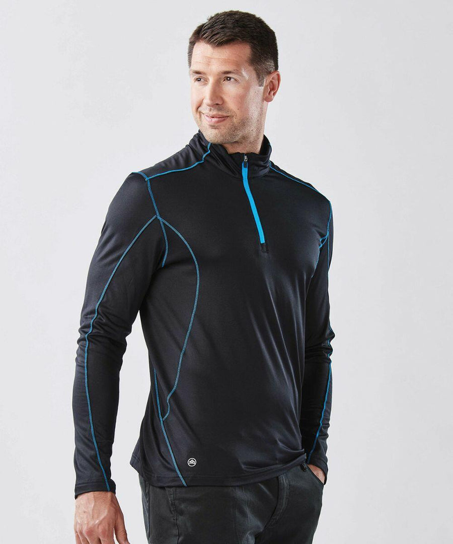 A man is wearing a Stormtech ST177 black long-sleeve athletic shirt with blue piping and a front zip. He stands with his hands in his pockets, looking to the side.