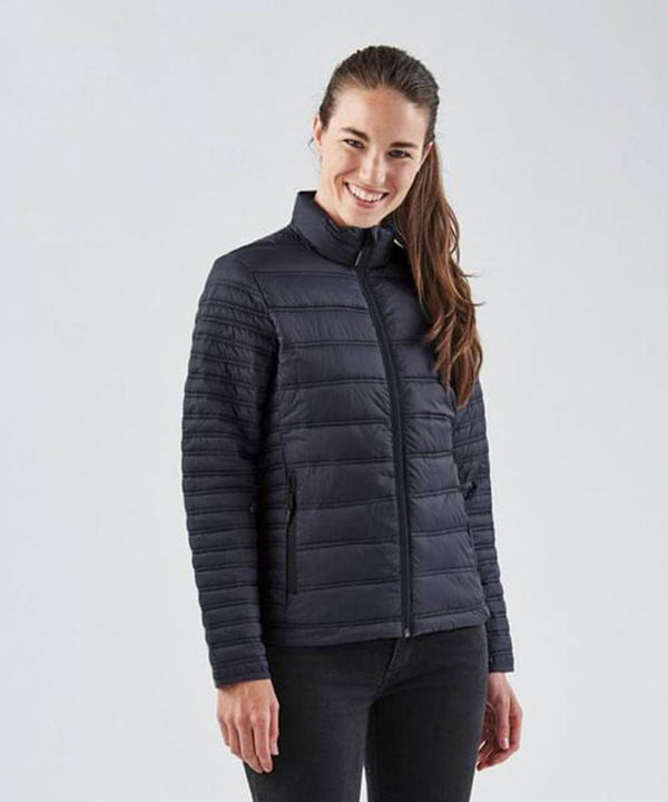 A woman with long brown hair stands smiling against a plain light background, wearing the Stormtech Women's Basecamp thermal jacket—a black quilted coat featuring a front zip and high collar.