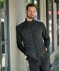A man with a beard wearing a black Stormtech ST195 stands with hands in pockets, leaning against a light-colored wall.