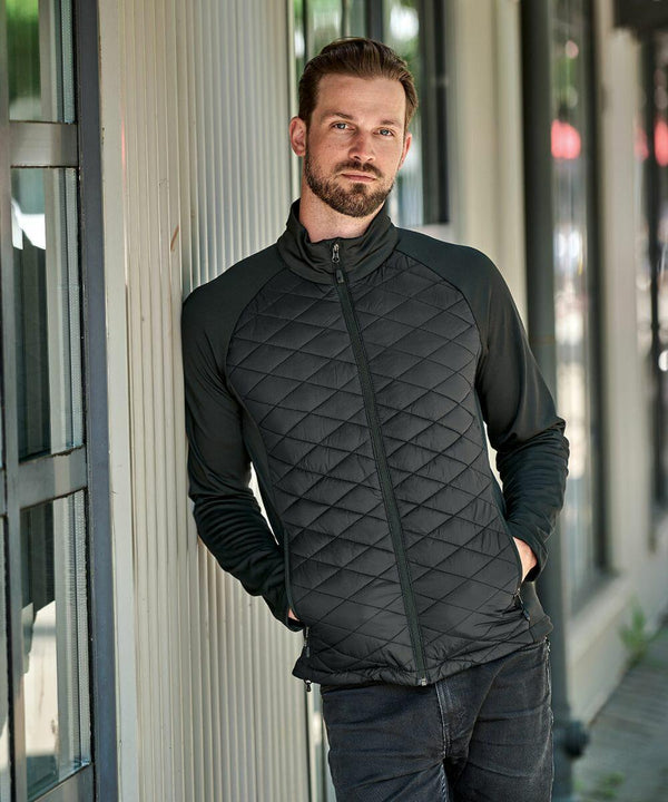 A man with a beard wearing a black Stormtech ST195 stands with hands in pockets, leaning against a light-colored wall.