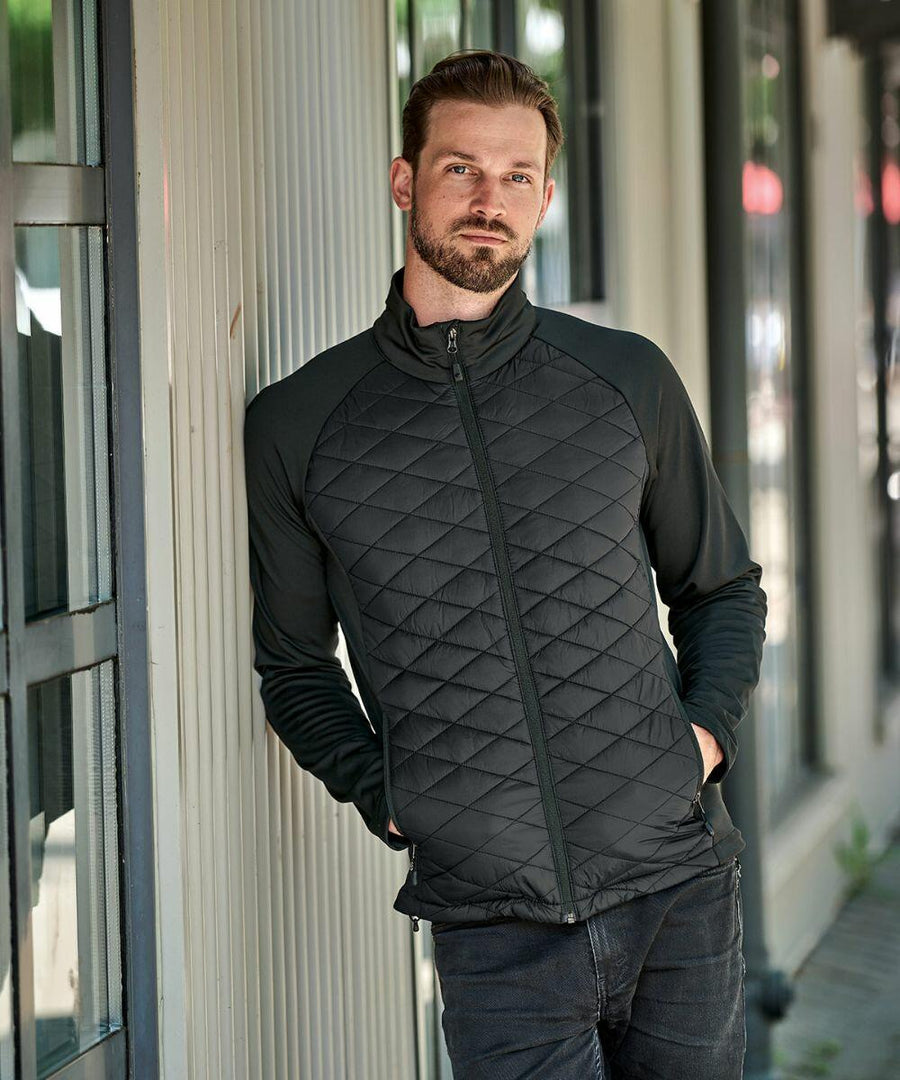 A man with a beard wearing a black Stormtech ST195 stands with hands in pockets, leaning against a light-colored wall.