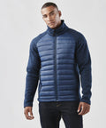 A man stands against a plain background, wearing a blue ST198 zip-up jacket with padded front and sleeves from Stormtech, paired with dark jeans.