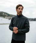A man with a beard stands outdoors near water, wearing a dark Stormtech ST200 zip-up jacket and looking to his left.