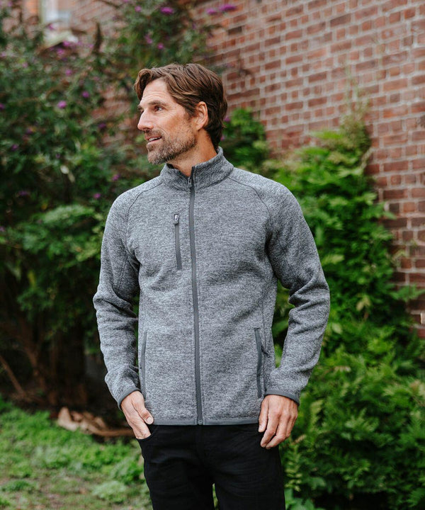 A man wearing a gray Stormtech ST204 zip-up jacket stands outside in front of a brick wall and greenery, looking to the side with his hands in the jacket pockets.