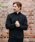 A man with short brown hair is standing in front of a brick wall, wearing a black Stormtech ST208 pullover and clasping his hands together. He is looking to his right.