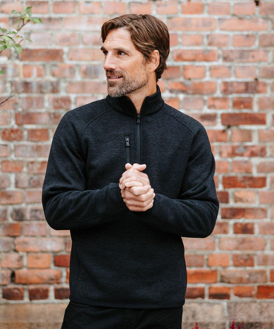 A man with short brown hair is standing in front of a brick wall, wearing a black Stormtech ST208 pullover and clasping his hands together. He is looking to his right.