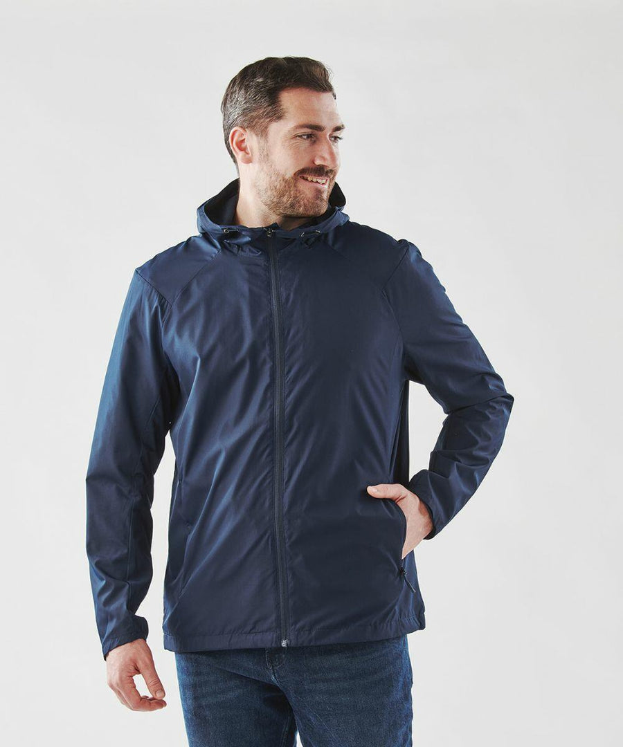 A man wearing a blue Stormtech ST210 hooded jacket and blue jeans stands with his left hand in his pocket, looking slightly to his right.