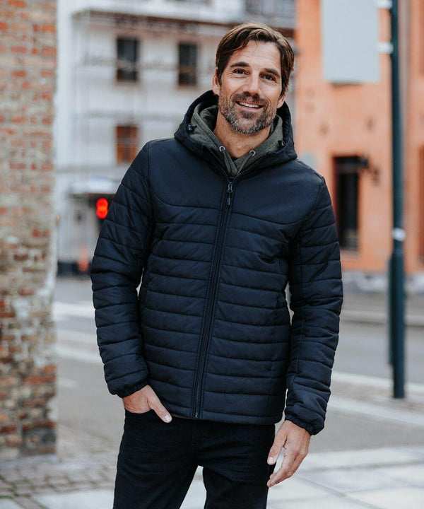 A man with a beard stands outside wearing a black Stormtech ST212 quilted jacket with a hood. He is smiling, and various buildings are visible in the background.