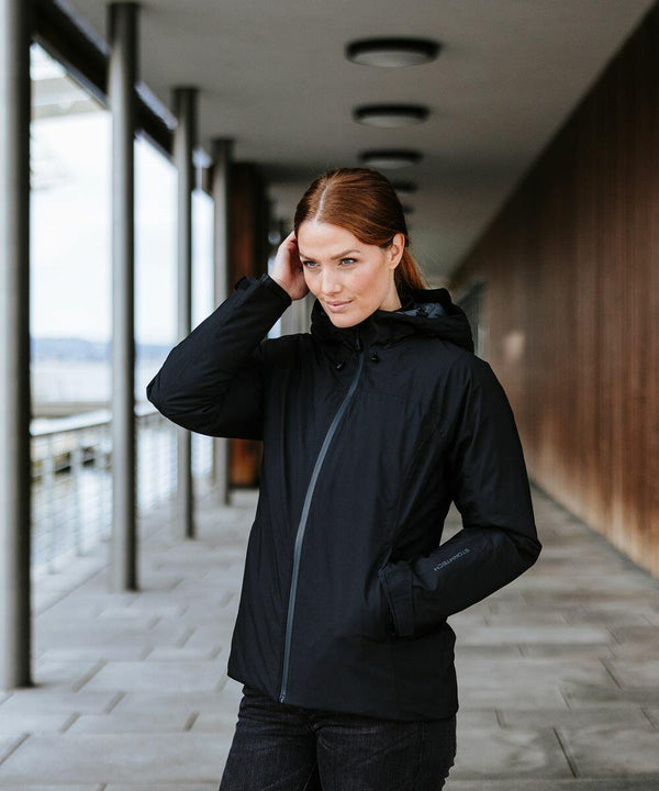 A woman with long red hair, wearing a black Stormtech ST215 jacket with her hand touching her hair, stands in a modern corridor with columns and glass panels.