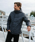 A person wearing a Stormtech ST218 stands casually on a bridge, looking to the side. The background shows an urban setting with trees and buildings.