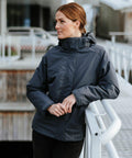 A person with brown hair stands outdoors wearing a dark gray Stormtech ST219 hooded jacket, leaning on a railing with a thoughtful expression.