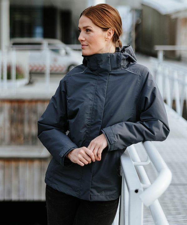 A person with brown hair stands outdoors wearing a dark gray Stormtech ST219 hooded jacket, leaning on a railing with a thoughtful expression.
