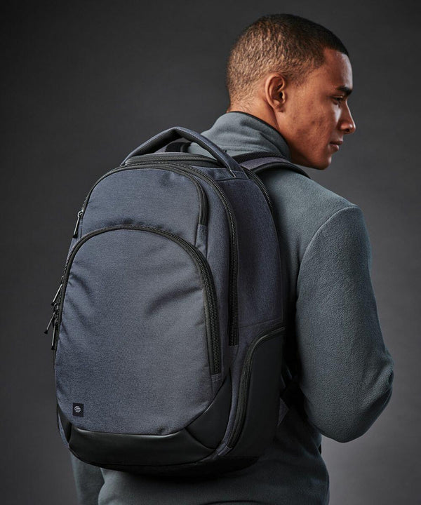 A person with short hair wearing a dark long-sleeve shirt and a large dark Stormtech ST221 backpack stands against a dark background, looking slightly to the right.