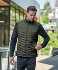 Man wearing a green Stormtech ST223 quilted vest over a black long-sleeve shirt while walking outdoors near a modern building.