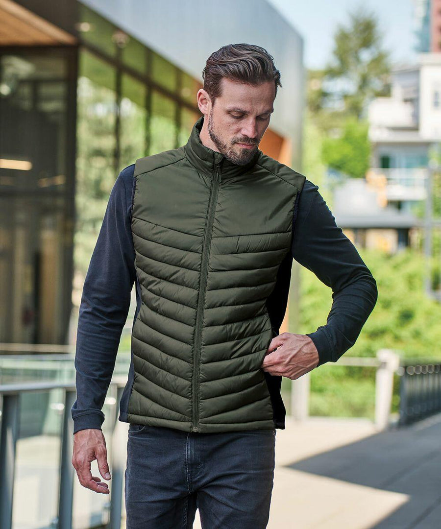 Man wearing a green Stormtech ST223 quilted vest over a black long-sleeve shirt while walking outdoors near a modern building.