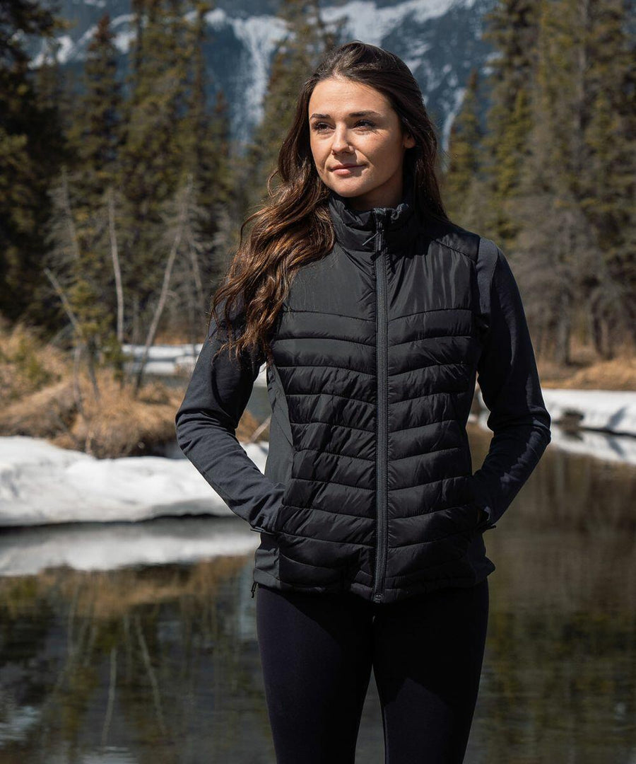 A woman stands outdoors wearing a black ST224 puffer vest and long-sleeve shirt by Stormtech. She has her hands in her pockets, with a forest and snowy mountains in the background.