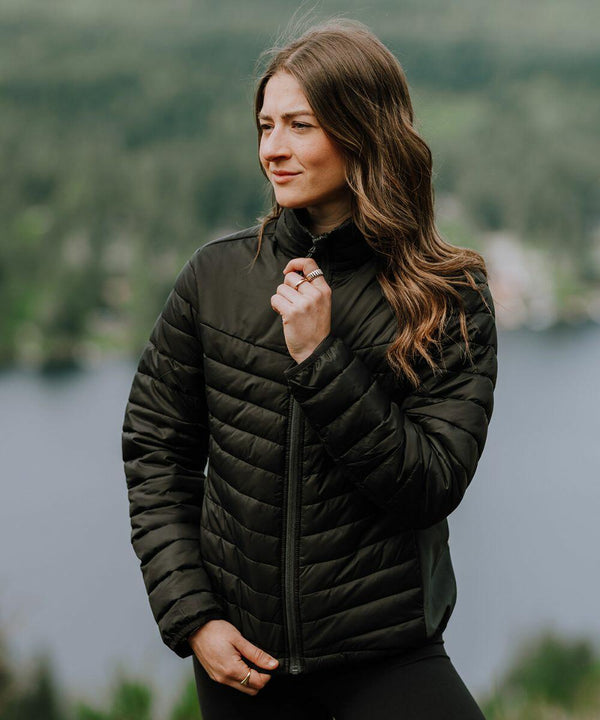 A woman with long hair wearing a black Stormtech ST226 jacket stands outdoors near a body of water with trees in the background.