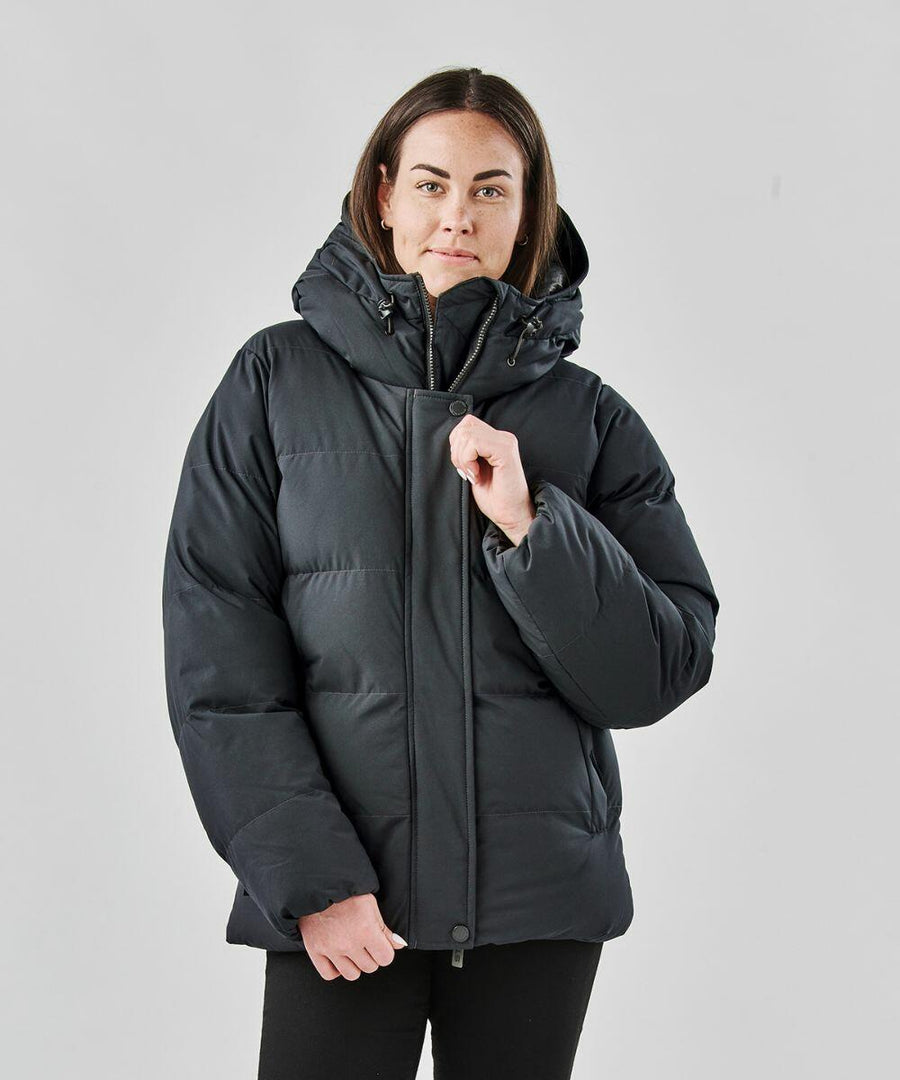 A person wearing a black Stormtech ST228 puffer jacket with the hood up, standing against a plain gray background.