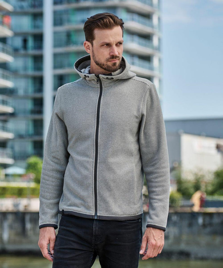 A man with short hair and a beard is wearing a gray Stormtech ST229 hooded jacket. He is standing outdoors with a modern building in the background.