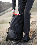 Person in black clothing and boots adjusts contents in a large black Stormtech ST234 backpack outdoors, with rocky and grassy terrain in the background.