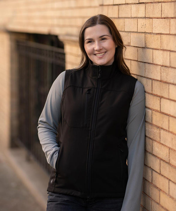 A woman with long brown hair stands outdoors against a brick wall, smiling at the camera. She wears a light grey long-sleeve shirt and a black Stormtech Women's Cascades softshell gilet.