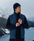 A person wearing a Stormtech Cascadia thermal gilet, blue beanie, and dark jacket stands outdoors by snowy mountains and a lake, looking to the side with hands near the jacket zip.