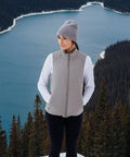 A woman in a Stormtech Women’s Cascadia thermal gilet, grey beanie, and white long-sleeved shirt stands on a snowy hilltop overlooking a lake bordered by evergreen trees.