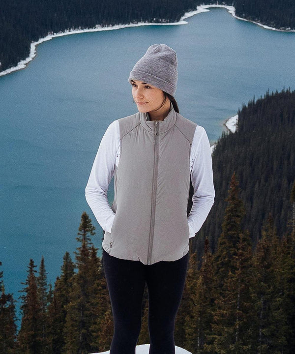 A woman in a Stormtech Women’s Cascadia thermal gilet, grey beanie, and white long-sleeved shirt stands on a snowy hilltop overlooking a lake bordered by evergreen trees.