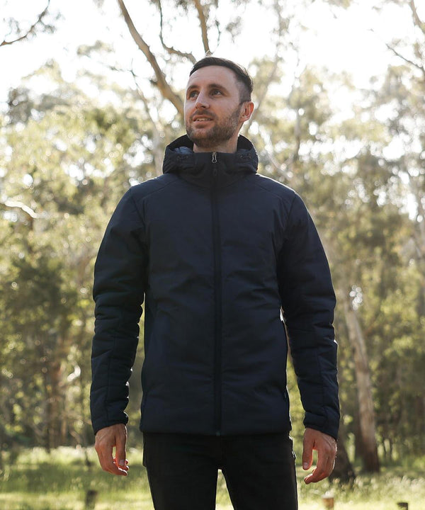 A man wearing the Stormtech Cascadia thermal jacket stands outdoors in a sunlit forest, looking slightly upward with trees and greenery in the background.