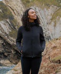 A woman with curly hair stands outdoors, smiling and looking up, hands in the pockets of her black Stormtech Women's Cascadia thermal jacket. Rocky cliffs and greenery form the backdrop.