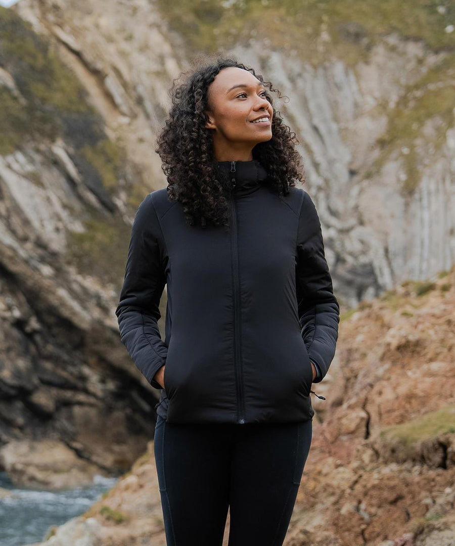 A woman with curly hair stands outdoors, smiling and looking up, hands in the pockets of her black Stormtech Women's Cascadia thermal jacket. Rocky cliffs and greenery form the backdrop.