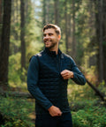 A man smiles as he zips up a Stormtech Appalachian thermal softshell gilet over a long-sleeve shirt in a sunlit forest filled with tall trees and green foliage.