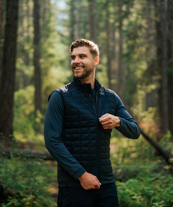 A man smiles as he zips up a Stormtech Appalachian thermal softshell gilet over a long-sleeve shirt in a sunlit forest filled with tall trees and green foliage.