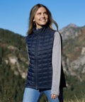 A woman wears the Stormtech Women's Appalachian thermal softshell gilet in navy over a grey long-sleeve shirt whilst standing outdoors with mountains and trees behind her, smiling and looking slightly to the side.