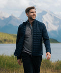 A man with short hair and a beard is walking by a lake, wearing the Stormtech Appalachian thermal softshell jacket. Mountains and trees are visible in the background beneath a partly cloudy sky.