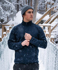 A man in a grey beanie and Stormtech Yosemite full-zip fleece jacket stands on a snowy bridge amid falling snow and trees, looking to the side with one hand touching his collar.